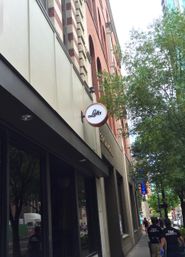 Downtown sidewalk beneath a red-brick multi-story building with a round storefront sign, leafy street trees and pedestrians walking past shops.
