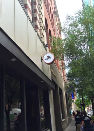 Downtown sidewalk beneath a red-brick multi-story building with a round storefront sign, leafy street trees and pedestrians walking past shops.