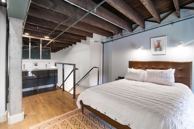 Cozy industrial loft bedroom with exposed dark wooden ceiling beams, leather tufted headboard, white ruffled bedding, patterned rug, hardwood floors and open-plan view of a modern double-sink vanity with subway tile and metal stair railing.