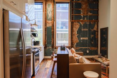 Open-plan urban loft apartment with exposed brick and distressed plaster wall, industrial kitchen with stainless refrigerator and stove, farmhouse sink, wood floors, bar seating and a small sofa facing a tall city-view window.