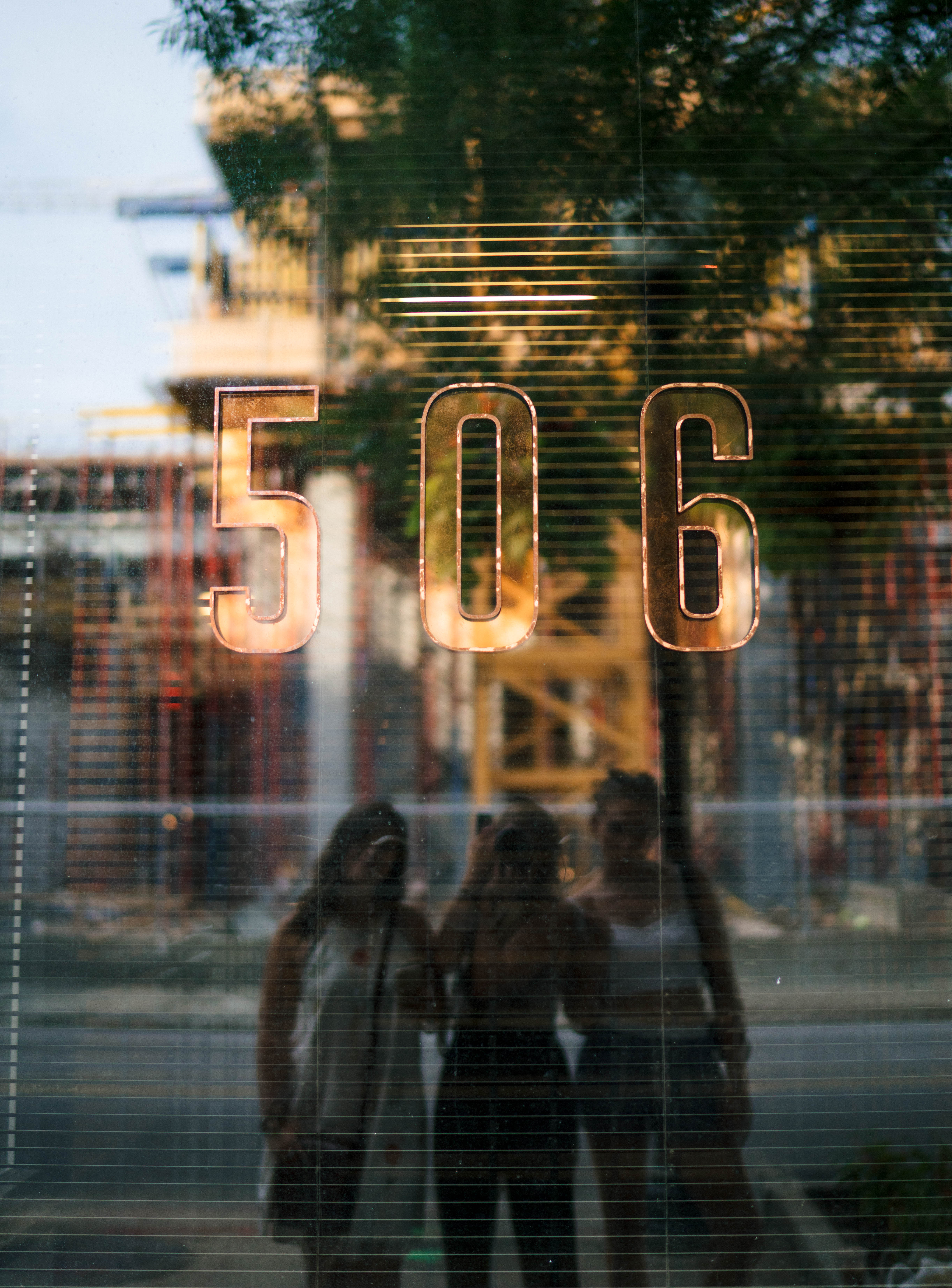 Shiny copper '506' on a reflective storefront window with an urban street and three silhouetted pedestrians reflected in the glass.