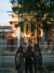 Shiny copper '506' on a reflective storefront window with an urban street and three silhouetted pedestrians reflected in the glass.