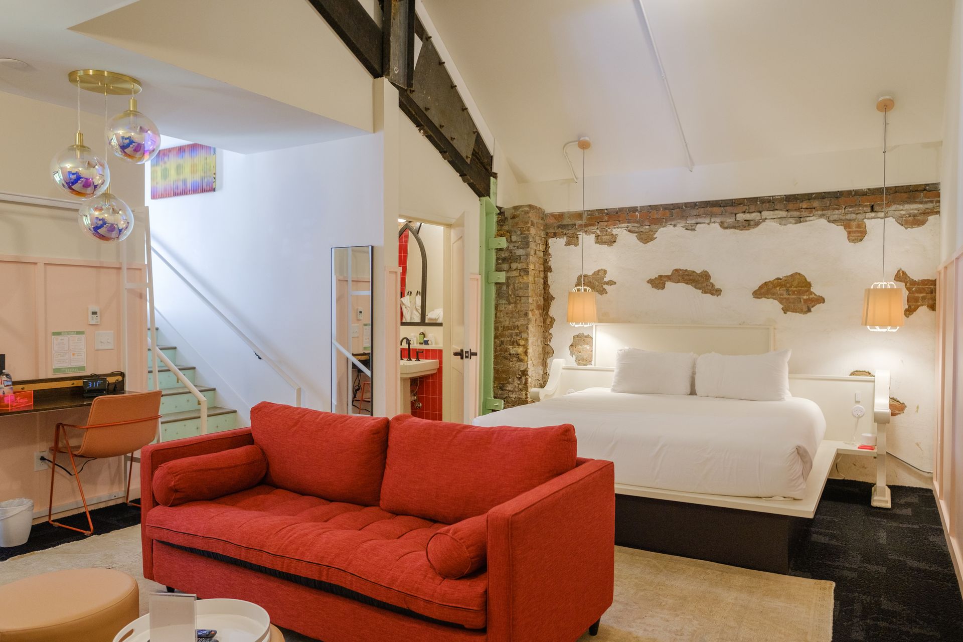 Cozy loft-style boutique hotel room with a king bed set against a partially exposed brick wall, two hanging pendant lamps, a vibrant red sofa in the foreground, small workspace, and stairs leading to an upper level with a peek of a red-tiled bathroom.