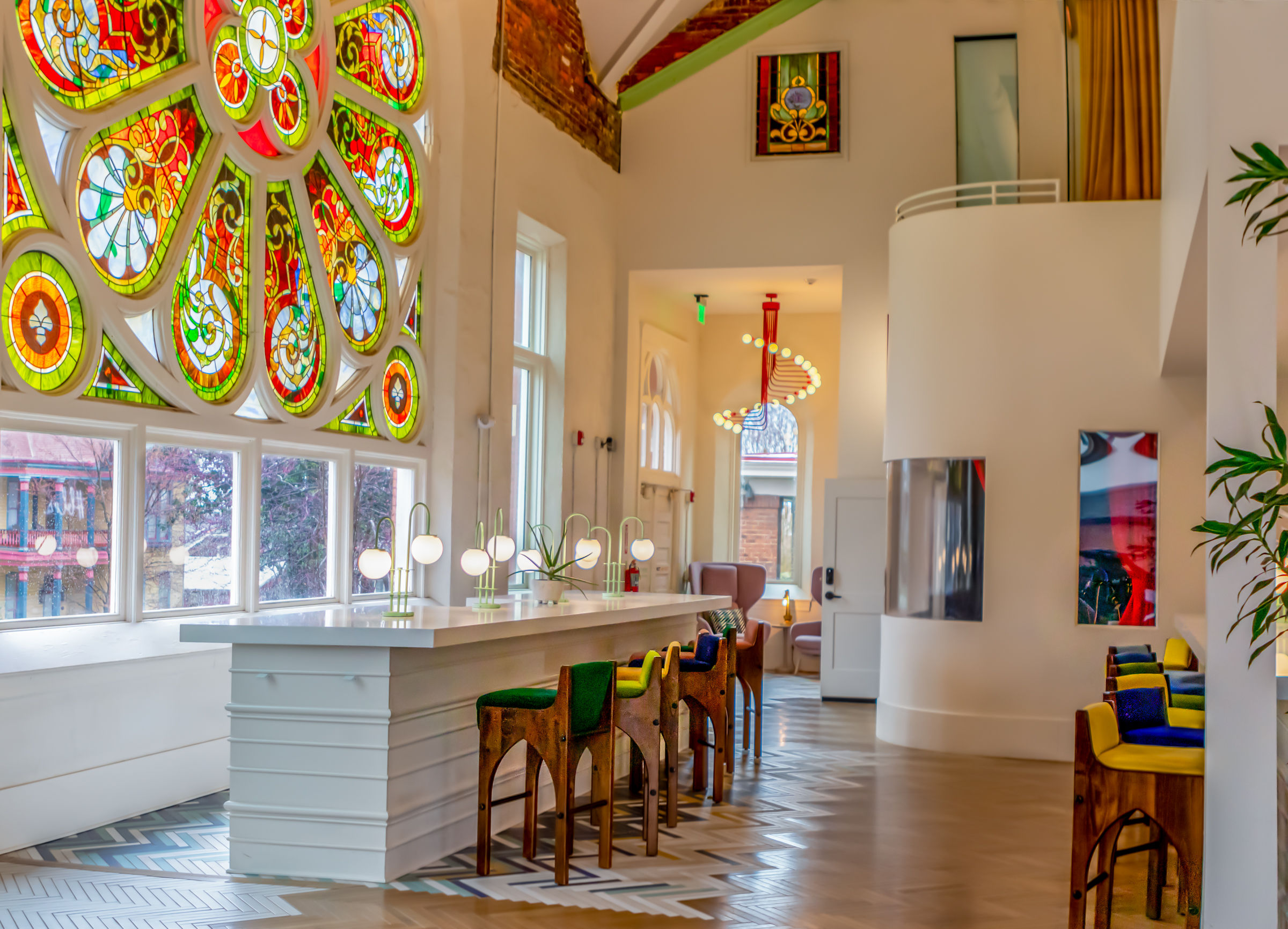 Sunlit cafe interior in a renovated historic building featuring a large colorful stained-glass rose window, long white bar with globe lamps, wooden high stools with green and blue cushions, patterned herringbone floor and modern pendant lights.
