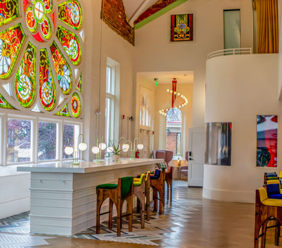 Sunlit cafe interior in a renovated historic building featuring a large colorful stained-glass rose window, long white bar with globe lamps, wooden high stools with green and blue cushions, patterned herringbone floor and modern pendant lights.
