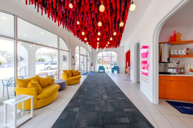 Sunlit boutique hotel lobby with dramatic red ribbon ceiling and hanging bulbs, cozy yellow circular sofas, blue armchairs by glass doors, and a cheerful orange refreshment nook.