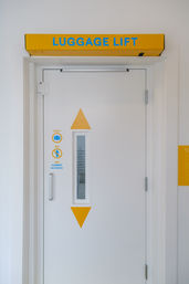 White luggage lift door in a bright corridor with a yellow header reading LUGGAGE LIFT, blue lettering, narrow vertical window, yellow arrow graphics and luggage-only icons.