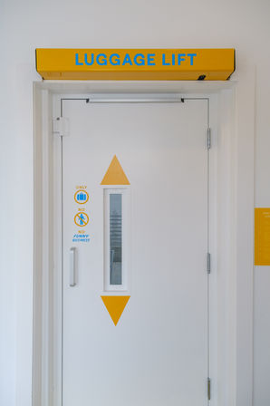 White luggage lift door in a bright corridor with a yellow header reading LUGGAGE LIFT, blue lettering, narrow vertical window, yellow arrow graphics and luggage-only icons.