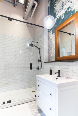 Urban loft bathroom with glass walk-in shower, white subway tile, matte-black shower fixtures, white vanity, leather-framed mirror and exposed industrial ceiling