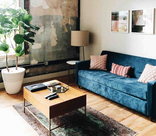 Sunlit loft-style living room with teal velvet sofa and patterned throw pillows, wooden coffee table on a vintage rug, floor lamp and large potted plant by an exposed-plaster wall.