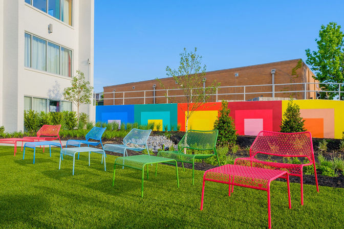 Bright urban courtyard with colorful metal lounge chairs on a manicured green lawn in front of a geometric rainbow mural and modern white building on a sunny day.