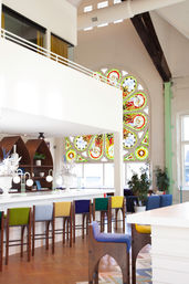 Sunlit loft-style cafe interior with a large ornate stained-glass window, white mezzanine, colorful upholstered bar stools, wooden floors, potted plants, and modern pendant lamps.