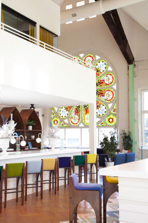 Sunlit loft-style cafe interior with a large ornate stained-glass window, white mezzanine, colorful upholstered bar stools, wooden floors, potted plants, and modern pendant lamps.