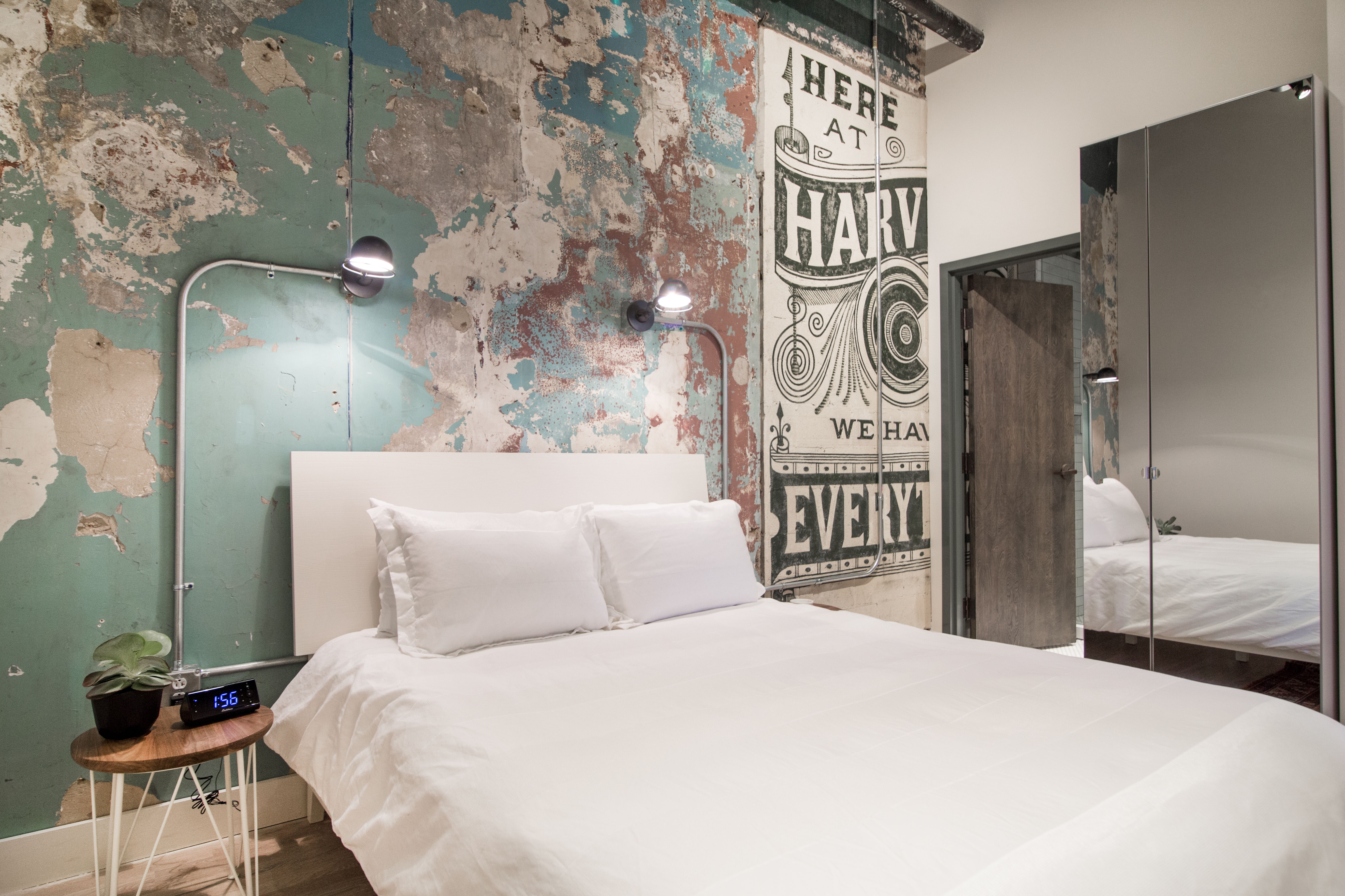 Urban loft bedroom with crisp white bedding against a distressed teal plaster wall and vintage-style painted mural, wall-mounted reading lamps, wooden side table with plant and digital alarm clock, and mirrored closet reflecting the bed.