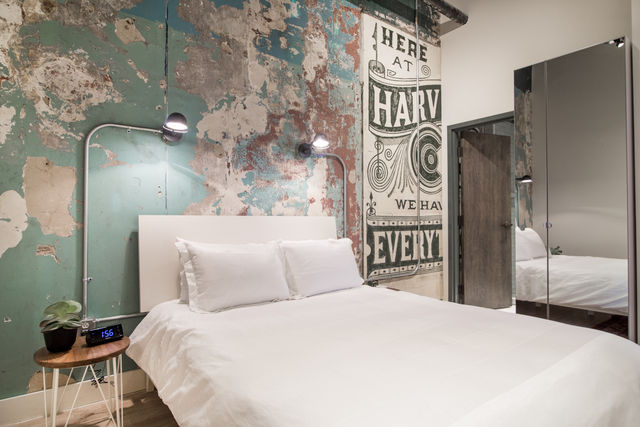 Urban loft bedroom with crisp white bedding against a distressed teal plaster wall and vintage-style painted mural, wall-mounted reading lamps, wooden side table with plant and digital alarm clock, and mirrored closet reflecting the bed.