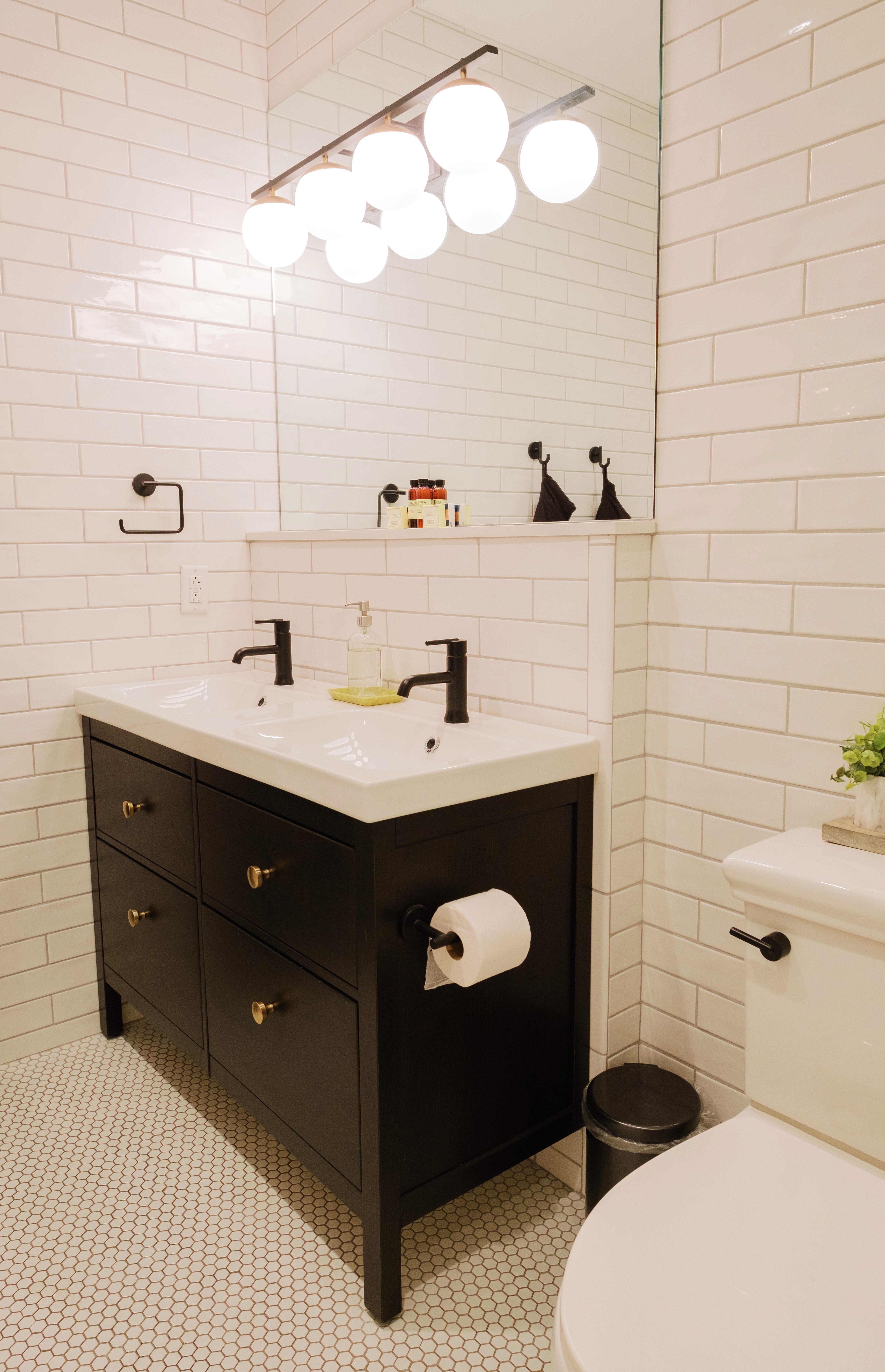 Bright modern bathroom with white subway tile and hexagon floor, sleek black double-sink vanity, matte black faucets, and globe vanity lights.
