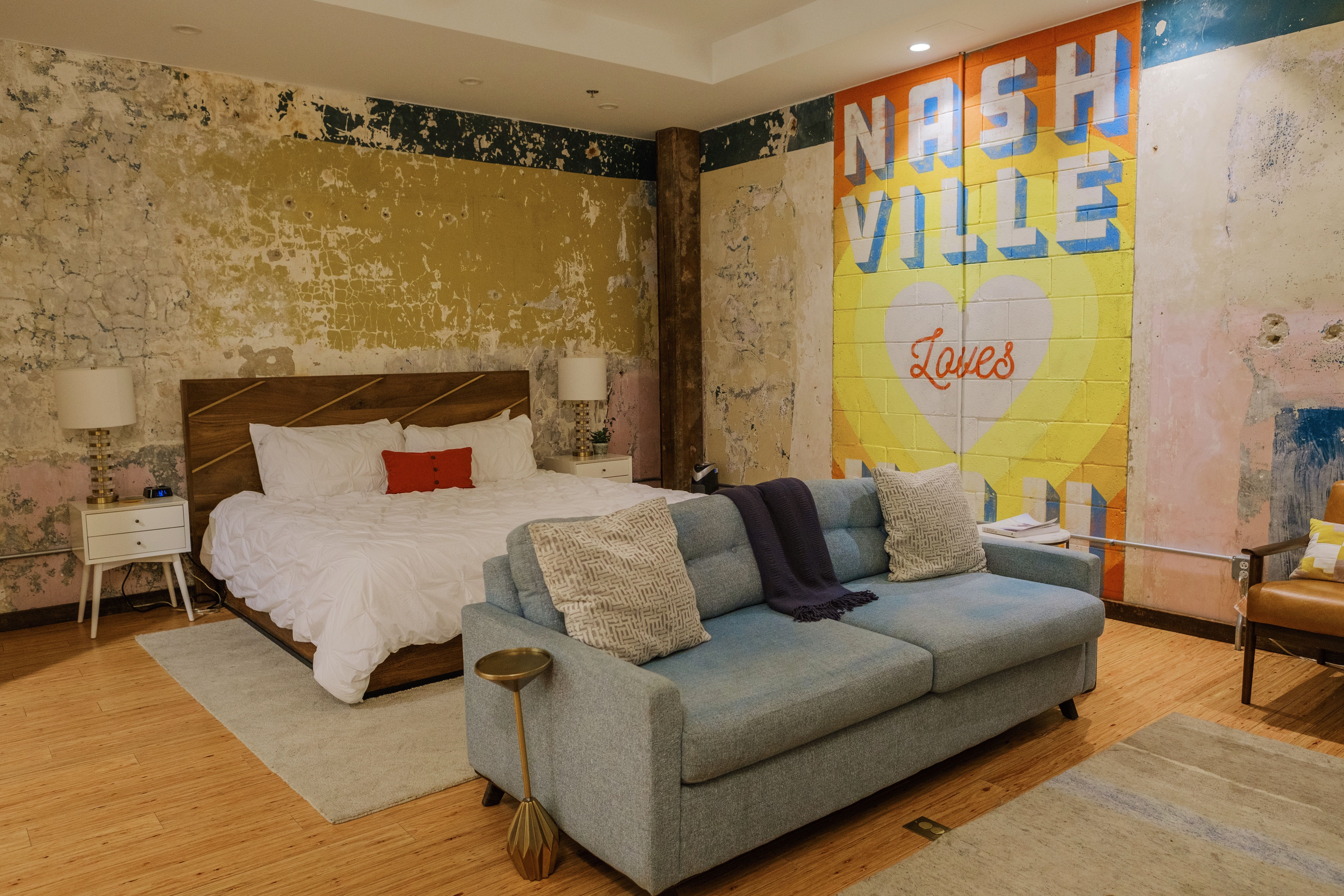 Stylish Nashville loft room with distressed plaster walls and bright 'Nashville Loves' mural, white bed, blue sofa, and hardwood floors