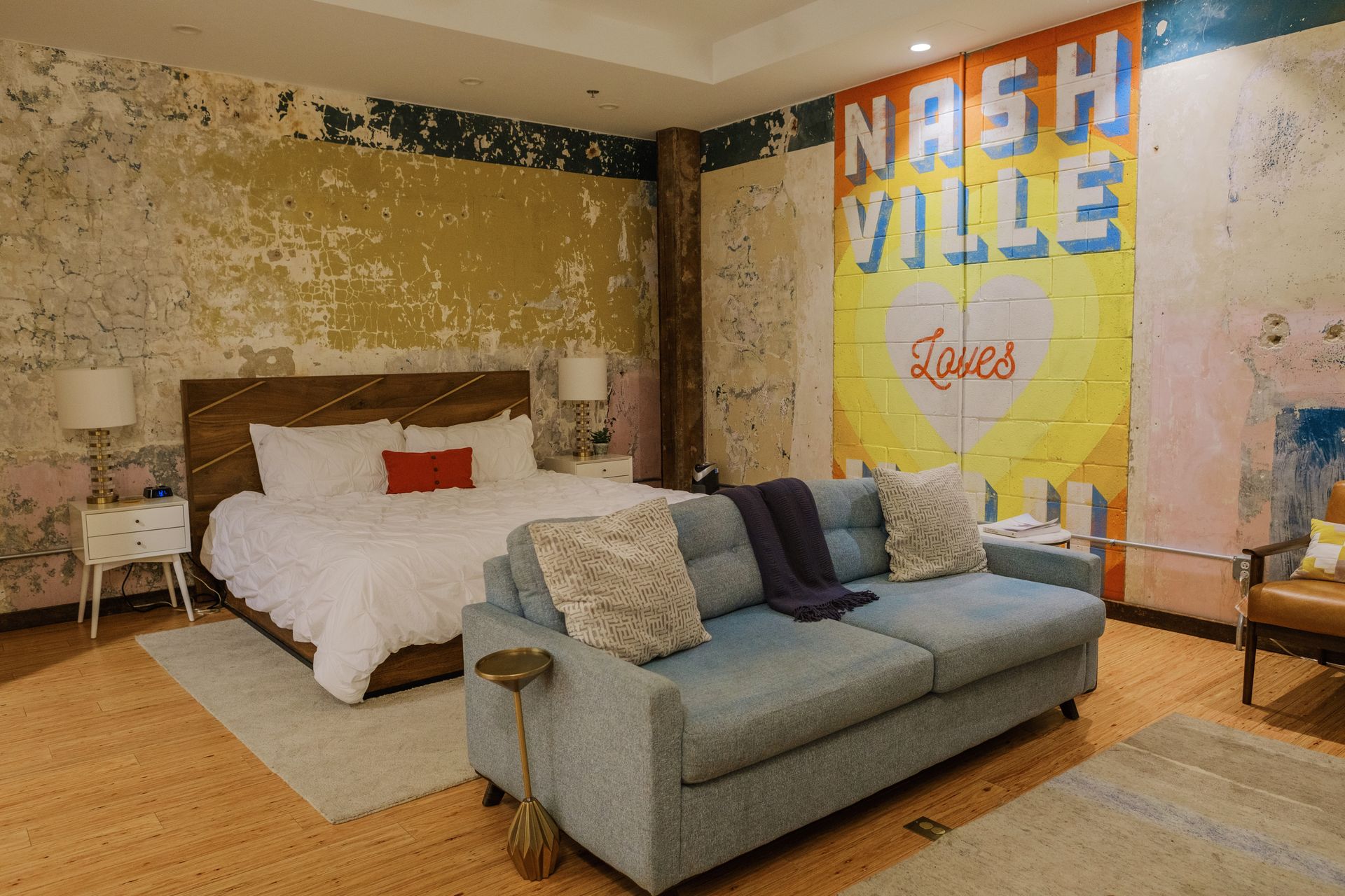 Stylish Nashville loft room with distressed plaster walls and bright 'Nashville Loves' mural, white bed, blue sofa, and hardwood floors