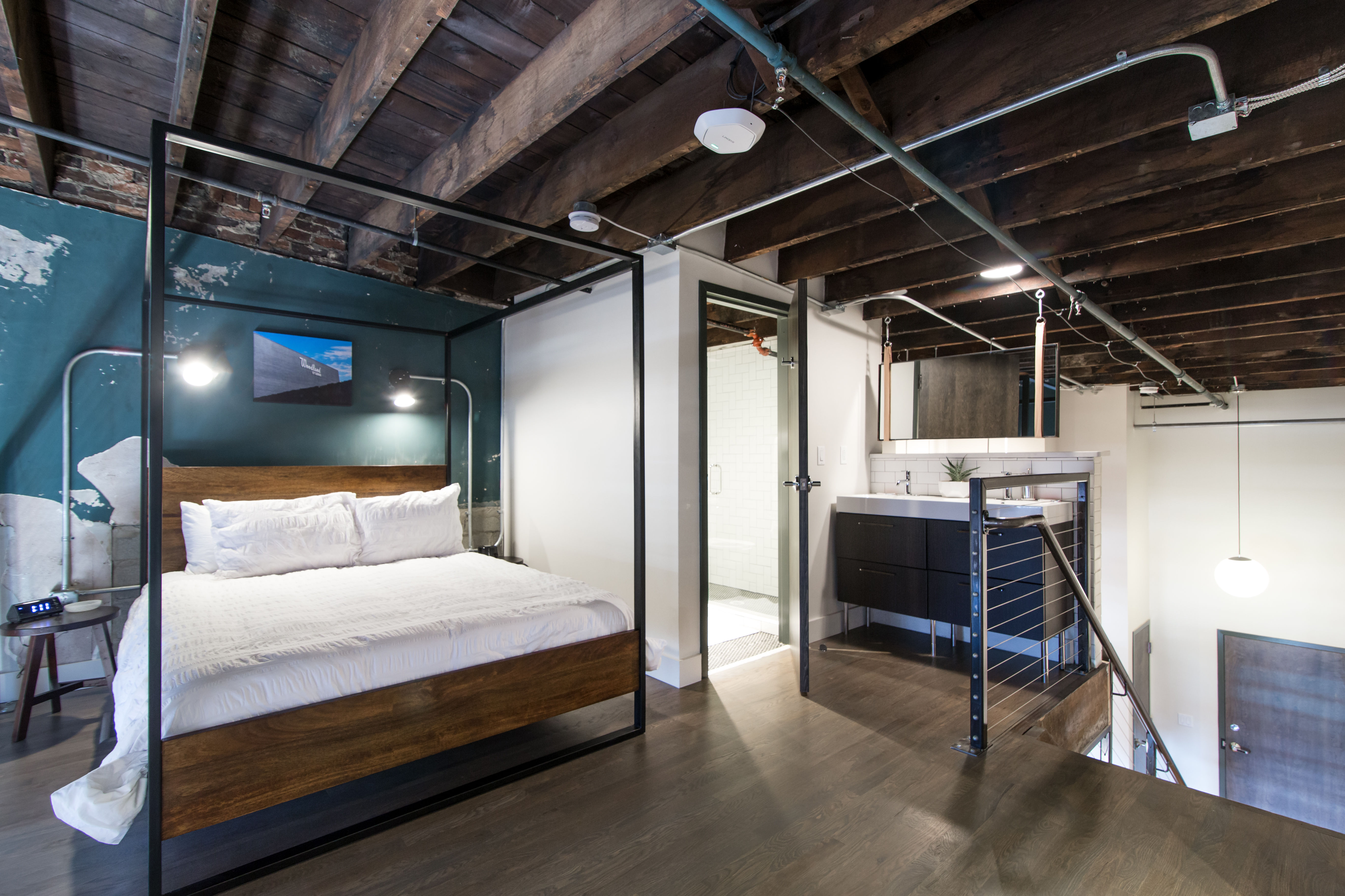 Industrial urban loft bedroom with exposed dark wooden beams, four-poster wood-and-metal bed dressed in white linens, teal accent wall, hardwood floors, open bathroom with modern vanity and glass shower, metal stair railing and hanging globe light.