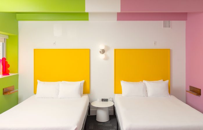 Bright, colorful hotel room with two beds, bold yellow headboards, crisp white linens, playful pink and green accent walls, and a central nightstand with clock and wall sconce.