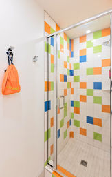 Bright modern glass-enclosed walk-in shower with playful orange, green, blue and white checkerboard tiles, tiled floor drain and an orange drawstring bag hanging on the wall.