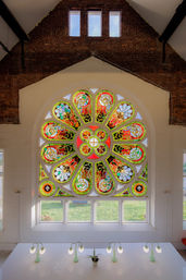 Vibrant circular stained-glass rose window with red, green and gold floral motifs in a brick vaulted interior, backlighting a white workspace with mint-green desk lamps and a small potted plant.