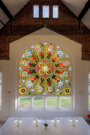 Vibrant circular stained-glass rose window with red, green and gold floral motifs in a brick vaulted interior, backlighting a white workspace with mint-green desk lamps and a small potted plant.