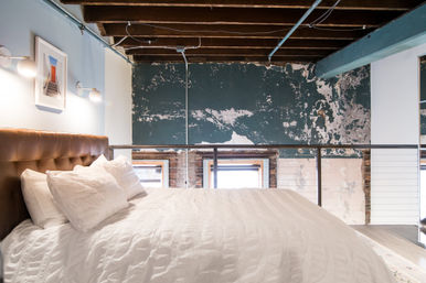 Cozy urban loft bedroom with white duvet and pillows, tufted leather headboard, exposed wooden ceiling beams, metal railing and distressed teal plaster wall.