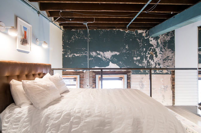 Cozy urban loft bedroom with white duvet and pillows, tufted leather headboard, exposed wooden ceiling beams, metal railing and distressed teal plaster wall.