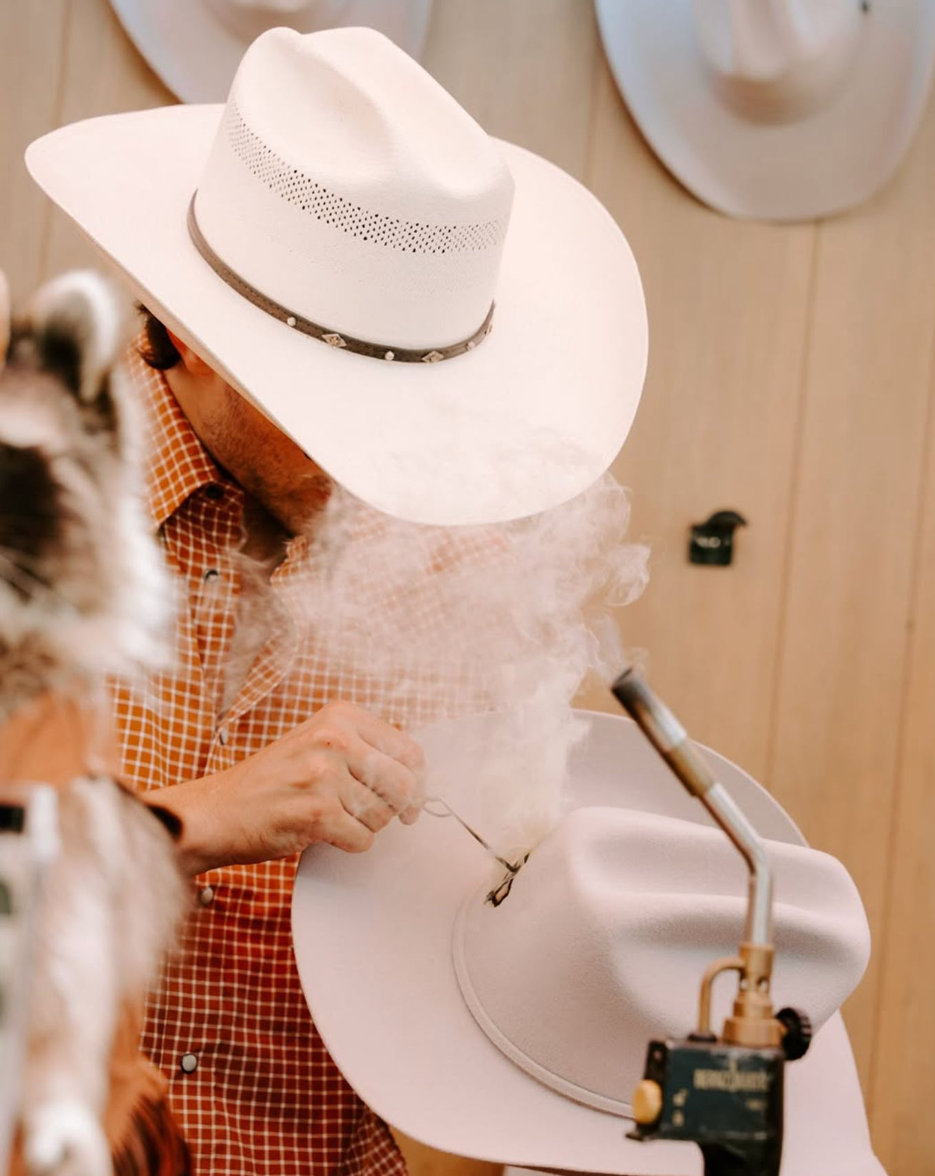 Hat maker shaping a white cowboy hat with a torch, smoke curling around the brim in a rustic western hat workshop.