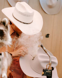 Hat maker shaping a white cowboy hat with a torch, smoke curling around the brim in a rustic western hat workshop.