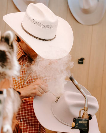 Hat maker shaping a white cowboy hat with a torch, smoke curling around the brim in a rustic western hat workshop.