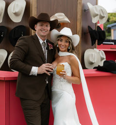 Smiling newlyweds in cowboy hats toast with cocktails at a western-themed wedding bar, red counter and a wall display of white and black cowboy hats