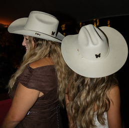 Two people with long wavy hair wearing white cowboy hats adorned with butterfly pins, seen from the side and back at an outdoor nighttime country-style gathering with warm string-light bokeh