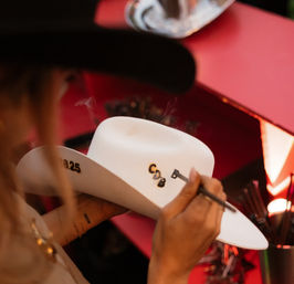 Close-up of an artisan customizing a white cowboy hat with a heated branding tool, pressing black initials into the brim as smoke rises over a red work surface.