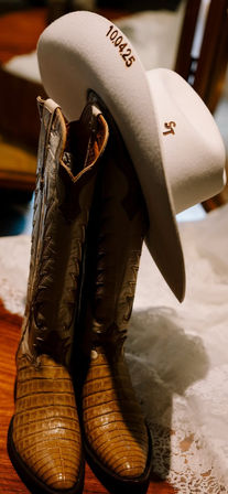 Tan crocodile-pattern cowboy boots paired with a white felt cowboy hat draped over them on a wooden floor and lace, hat stamped "10.04.25" — classic western accessories.