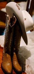 Tan crocodile-pattern cowboy boots paired with a white felt cowboy hat draped over them on a wooden floor and lace, hat stamped "10.04.25" — classic western accessories.