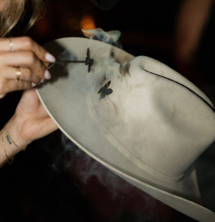 Close-up of hands using a small branding iron to add a smoky butterfly logo to a beige felt fedora