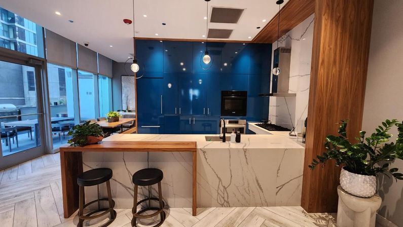 Sleek city apartment open-plan kitchen with glossy deep-blue cabinets, marble waterfall island and wood breakfast bar, two black stools, pendant lights, potted plants, and floor-to-ceiling windows opening to an outdoor patio.