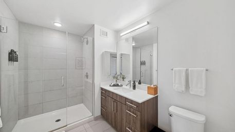 Bright spa-like modern bathroom with a frameless glass walk-in shower, wood vanity with white countertop and sink under a large mirror, towel rack and toilet.