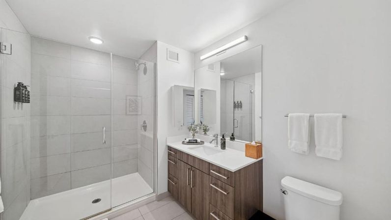 Bright spa-like modern bathroom with a frameless glass walk-in shower, wood vanity with white countertop and sink under a large mirror, towel rack and toilet.