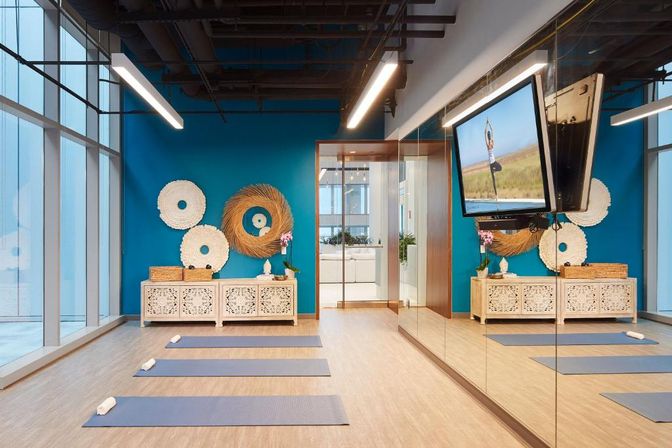 Bright modern yoga studio with floor-to-ceiling windows, blue accent wall and woven wall art; three blue mats with rolled towels on light wood floor, mirrored wall and TV showing a yoga pose.