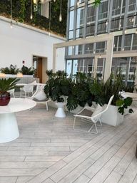 Modern sunlit atrium lounge in an urban high-rise with floor-to-ceiling glass, hanging filament pendant lights, white wicker chairs and tables, and abundant potted tropical plants with a vertical green wall.