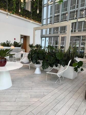 Modern sunlit atrium lounge in an urban high-rise with floor-to-ceiling glass, hanging filament pendant lights, white wicker chairs and tables, and abundant potted tropical plants with a vertical green wall.