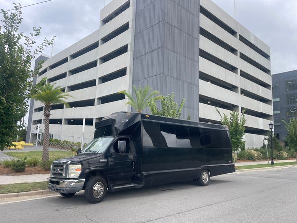 Black shuttle-style passenger van parked on an urban street beside palm trees in front of a multi-level concrete parking garage under an overcast sky.