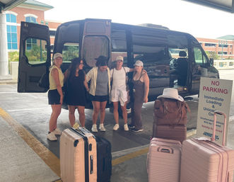 Group of five travelers posing by a black shuttle van under an airport pickup canopy with stacked pink and tan suitcases and a nearby “No Parking” sign