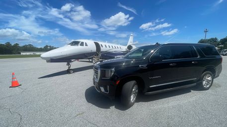 Black luxury SUV parked beside a white private jet on an airport tarmac under a bright blue sky with scattered clouds and an orange safety cone.