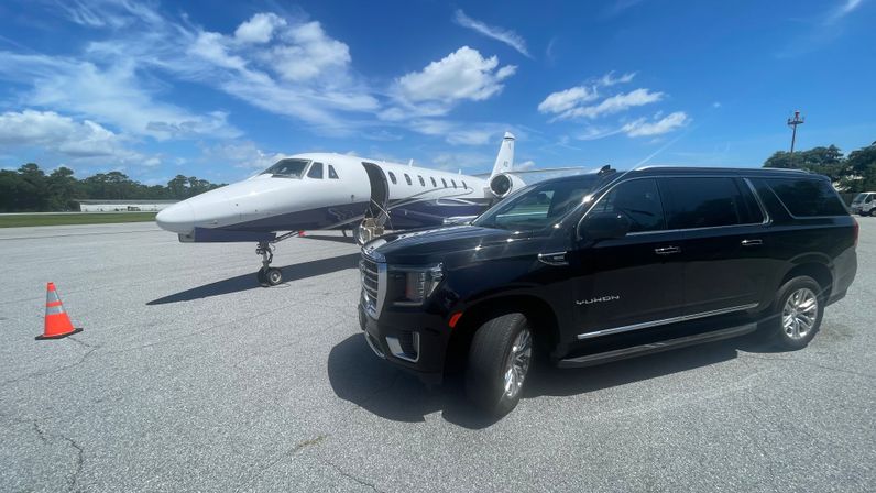 Black luxury SUV parked beside a white private jet on an airport tarmac under a bright blue sky with scattered clouds and an orange safety cone.