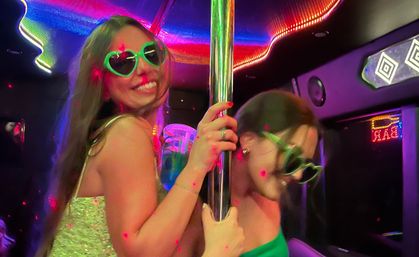 Two friends in a neon-lit party bus at night, wearing green heart-shaped sunglasses and smiling while holding a chrome dance pole under colorful LED lights