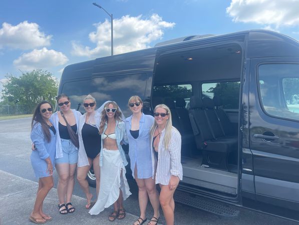 Six sun-ready women in swimsuits and light cover-ups posing by an open black passenger van in a sunny parking lot under blue skies.